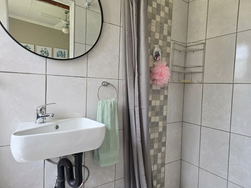 A compact bathroom featuring a wall-mounted basin with chrome fixtures, tiled shower area with mosaic accent, and a circular mirror.