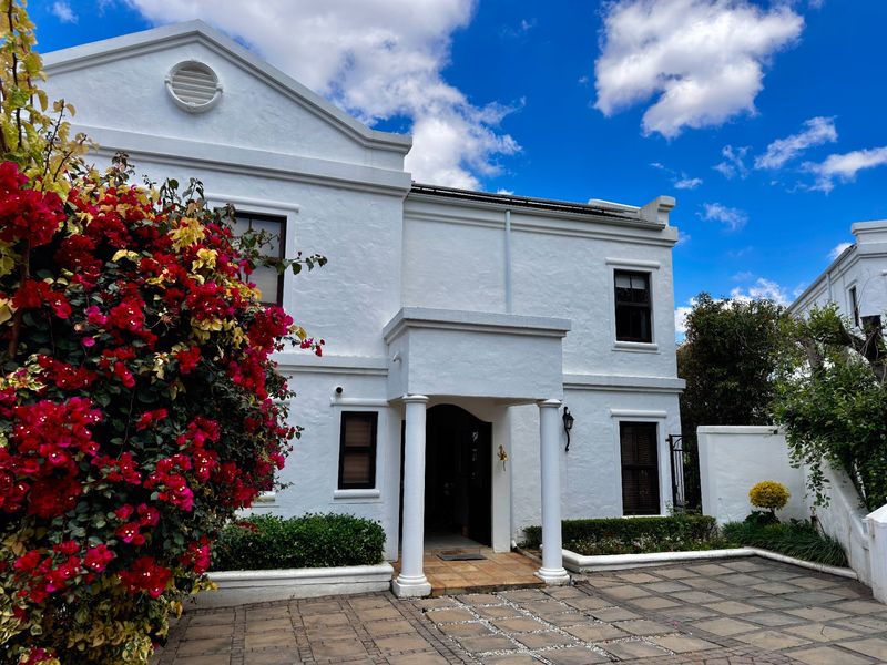 A well-maintained double-story home featuring a classic white facade, covered entrance with columns, and a paved driveway with established garden beds.