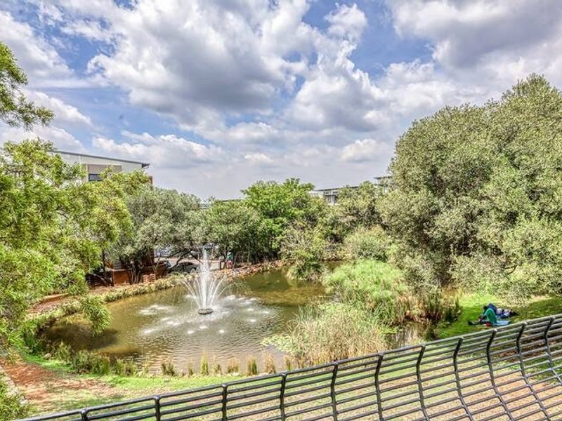 A scenic view overlooking a landscaped garden area featuring a central water fountain and mature trees, viewed from a balcony with a metal balustrade.