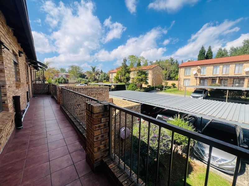 A spacious tiled balcony featuring a brick and metal balustrade, overlooking the complex parking area and surrounding residential grounds.