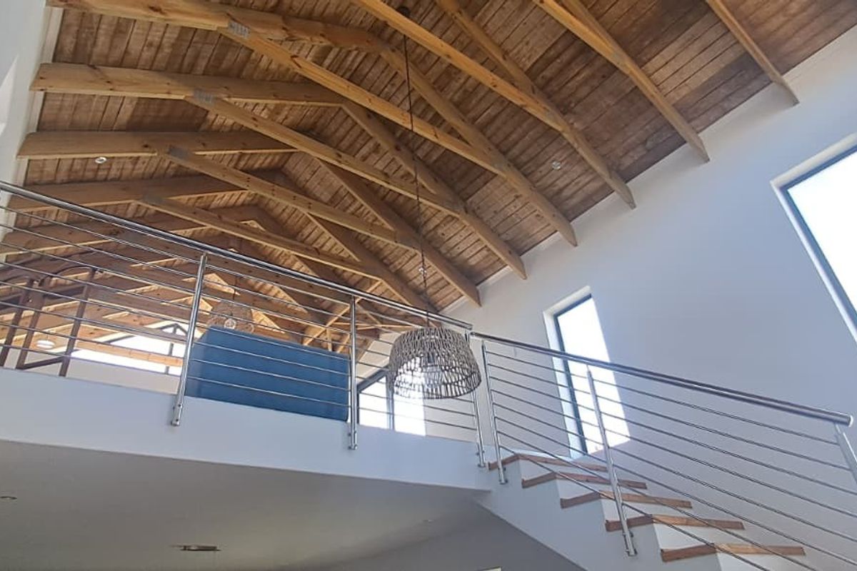 Spacious living area featuring a double-volume ceiling with exposed timber trusses, a mezzanine level, and a modern staircase with stainless steel balustrades.