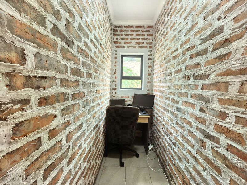 A compact home office featuring rustic exposed brick walls, tiled flooring, and a single window providing natural light.