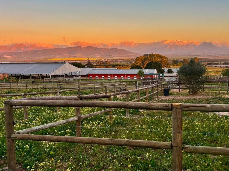 Scenic view of agricultural outbuildings and paddocks with timber fencing, set against a dramatic mountain backdrop at sunset.