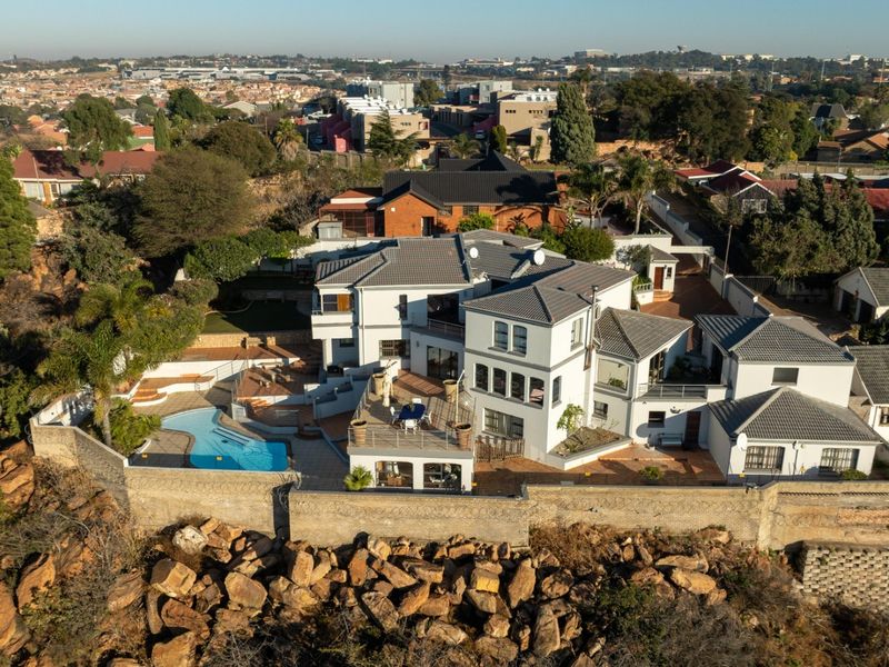Aerial view of a multi-level residential property featuring a swimming pool, expansive paved patio areas, and a tiled roof, situated on a rocky hillside.