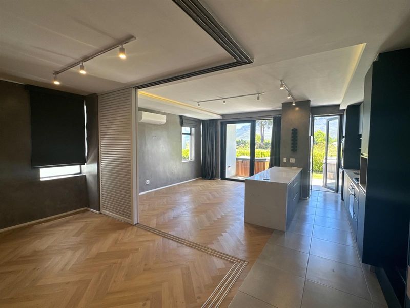 Spacious open-plan living and kitchen area featuring herringbone timber flooring, modern track lighting, and seamless access to an outdoor patio through sliding glass doors.