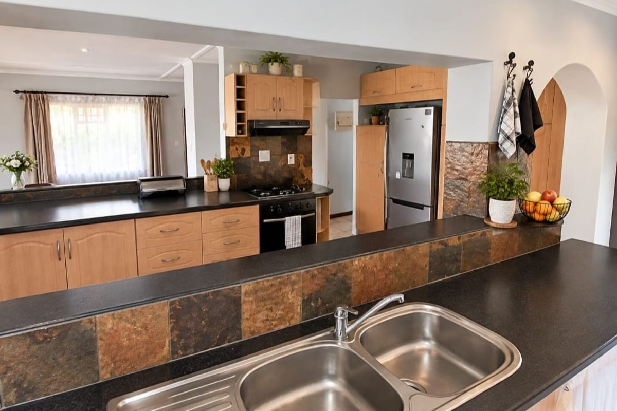 A functional kitchen featuring wooden cabinetry, stone tile splashbacks, dark laminate countertops, and a double bowl stainless steel sink with a breakfast bar pass-through.