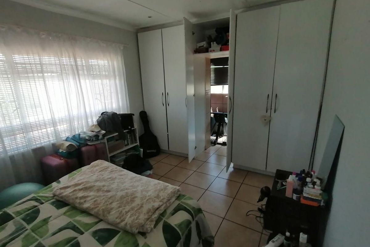 A bedroom featuring built-in white cupboards, tiled flooring, and large windows with blinds, offering a functional space with potential for cosmetic updates.