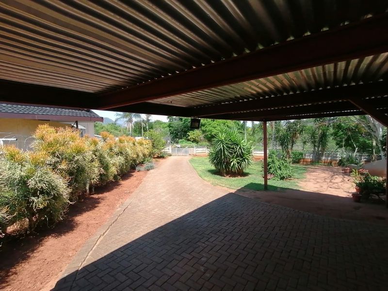 A covered outdoor carport area featuring a corrugated metal roof, steel support structure, and brick paving, overlooking a landscaped garden and driveway.