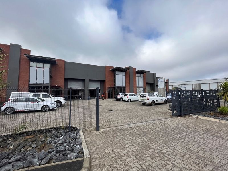 Front elevation of an industrial property featuring a paved driveway, secure gated entrance, and multiple roller shutter doors with modern brick and dark cladding.