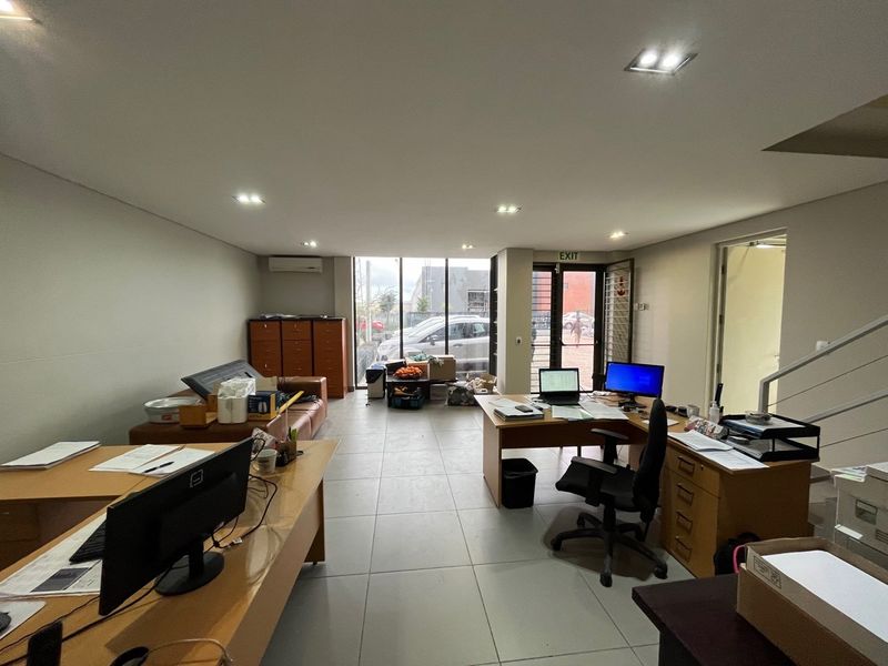 A functional open-plan office space featuring tiled flooring, recessed lighting, split-system air conditioning, and large windows providing natural light and exterior access.