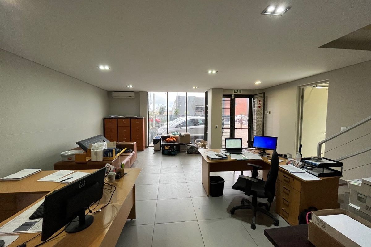 A functional open-plan office space featuring tiled flooring, recessed lighting, split-system air conditioning, and large windows providing natural light and exterior access.