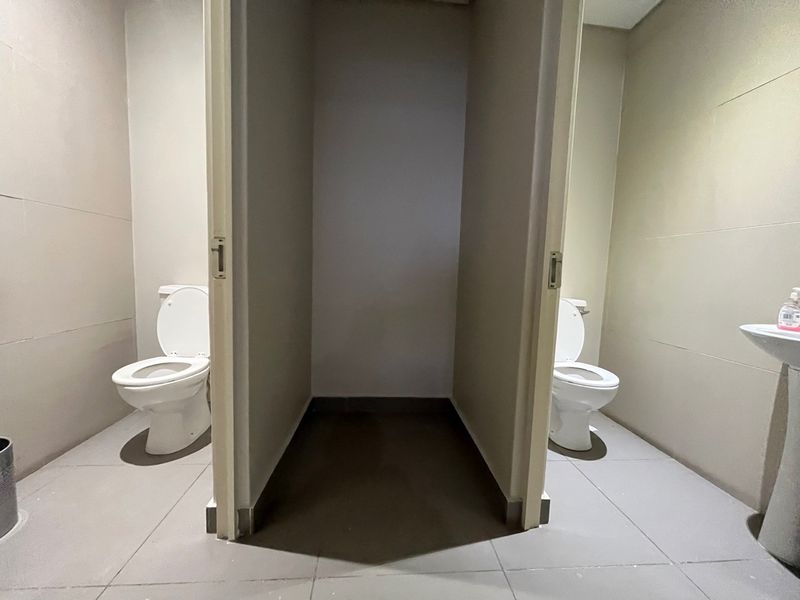 A functional industrial-style bathroom featuring two separate toilet cubicles and a pedestal basin with tiled flooring and neutral wall finishes.