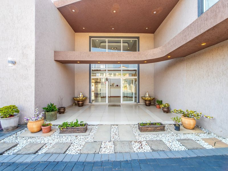 A modern, covered entrance featuring a glass-paneled doorway, tiled flooring, and a decorative stone-paved walkway leading to the main building facade.
