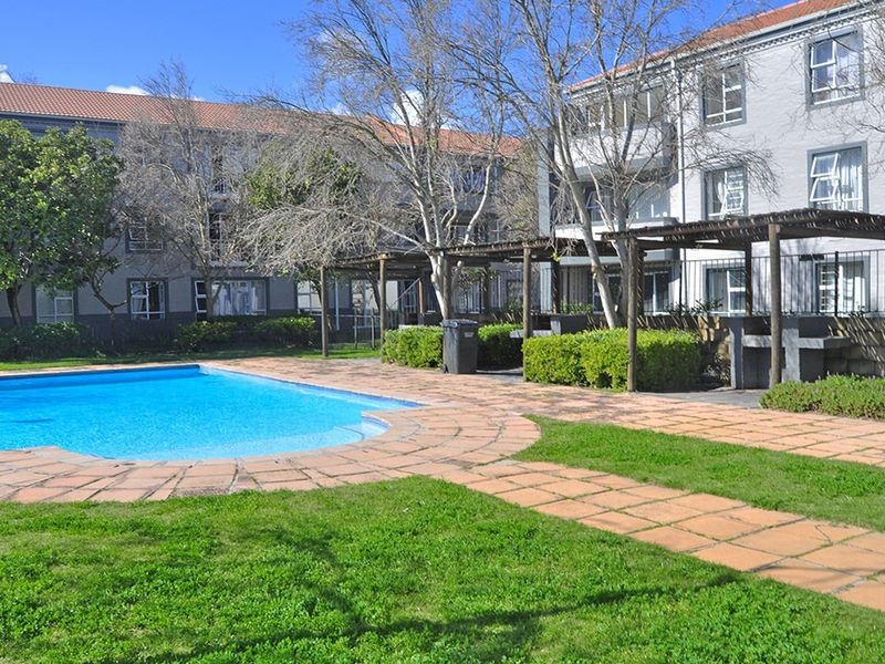 Communal apartment complex pool area featuring a swimming pool, paved surround, timber pergola, and built-in braai facilities set within a landscaped garden.