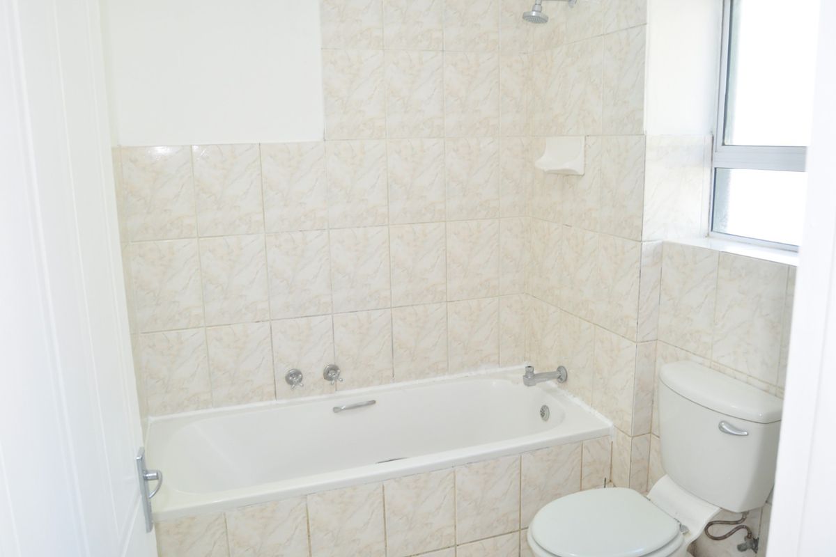 A dated bathroom featuring a built-in bathtub with shower head, toilet, and neutral patterned wall tiles.