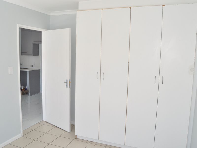 A bright, empty bedroom featuring built-in white cupboards, tiled flooring, and a view through to an adjacent kitchen area.