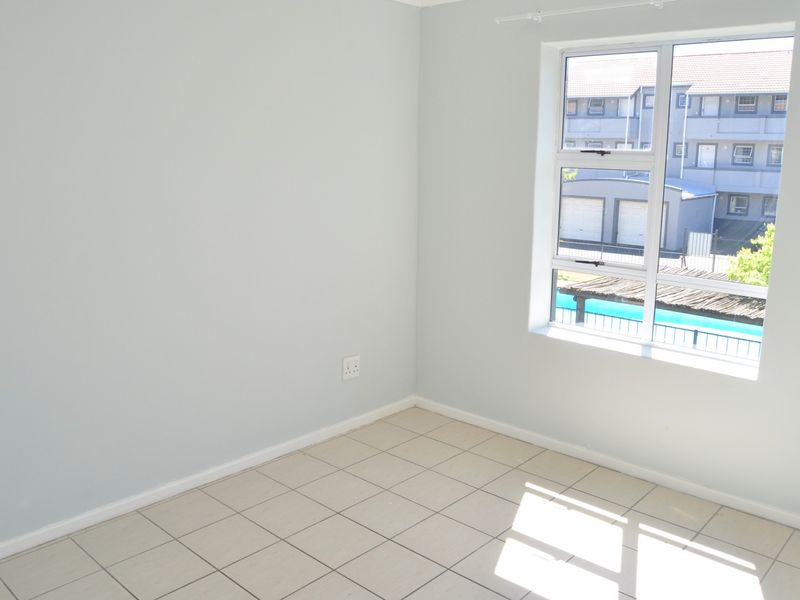 A bright, empty bedroom featuring neutral tiled flooring, light-colored walls, and a large window providing natural light and a view of the complex.