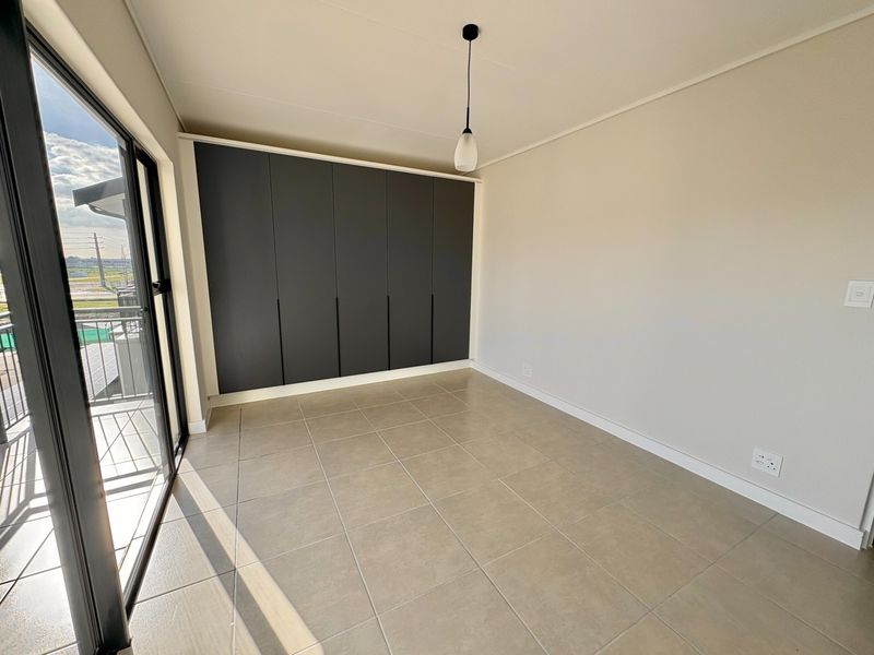 A bright, empty bedroom featuring modern dark built-in cupboards, neutral tiled flooring, and a sliding glass door leading to an outdoor balcony.