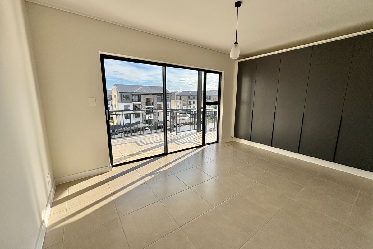 A bright, modern bedroom featuring neutral tiled floors, floor-to-ceiling built-in cupboards, and a large sliding door opening onto a private balcony.