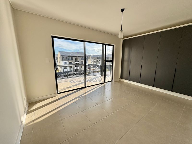 A bright, modern bedroom featuring neutral tiled floors, floor-to-ceiling built-in cupboards, and a large sliding door opening onto a private balcony.