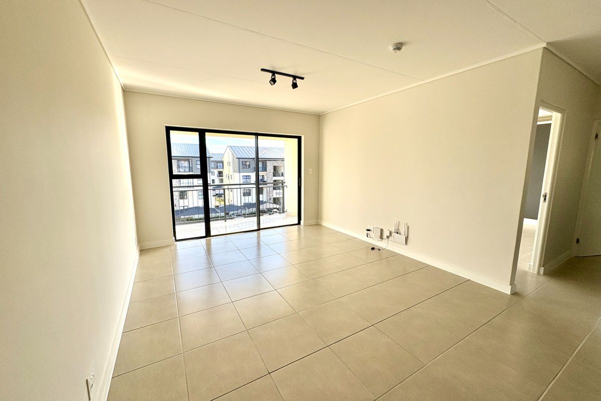 A bright, empty living room featuring neutral tiled flooring, modern track lighting, and a large sliding glass door leading to a balcony.