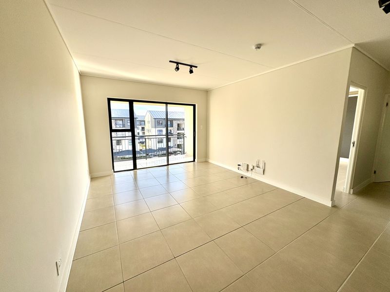 A bright, empty living room featuring neutral tiled flooring, modern track lighting, and a large sliding glass door leading to a balcony.