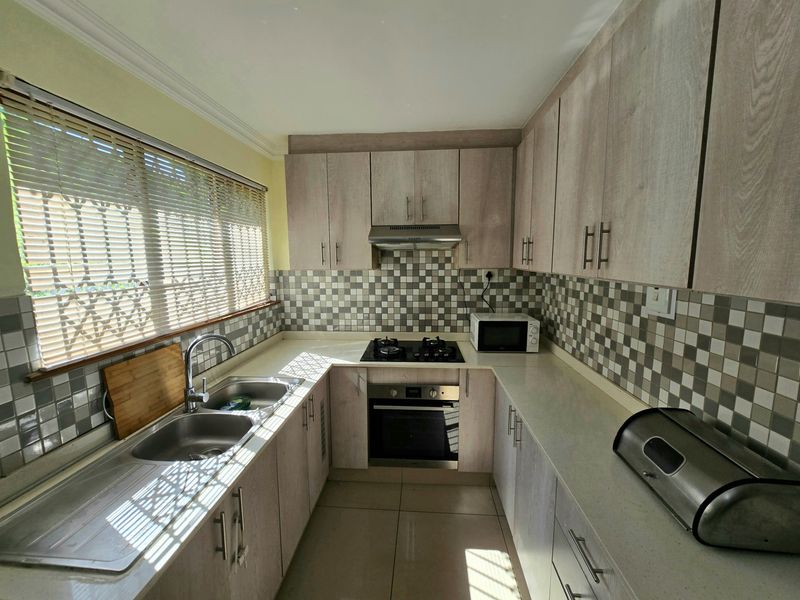 A functional kitchen featuring light wood-grain cabinetry, stone countertops, a gas hob with extractor, and a tiled backsplash with a double sink.