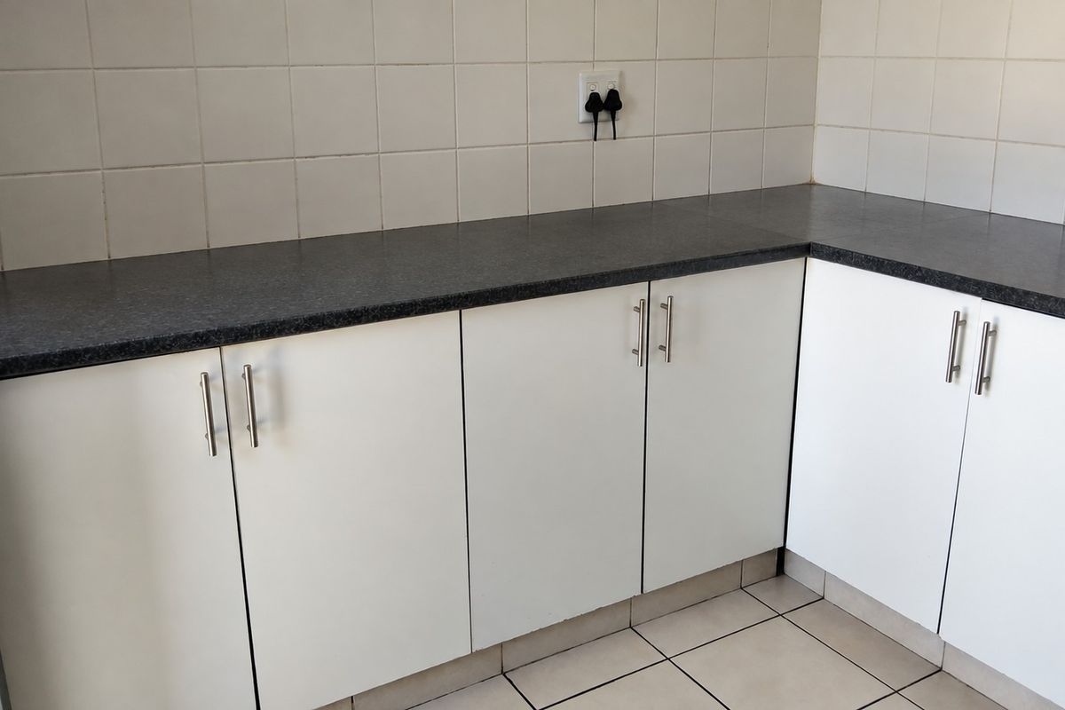 A clean, functional kitchen corner featuring white cabinetry, dark laminate countertops, and neutral ceramic wall and floor tiling.