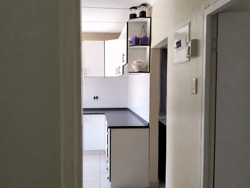 A compact kitchen featuring white built-in cabinetry, dark laminate countertops, and tiled flooring, viewed from an adjacent hallway.