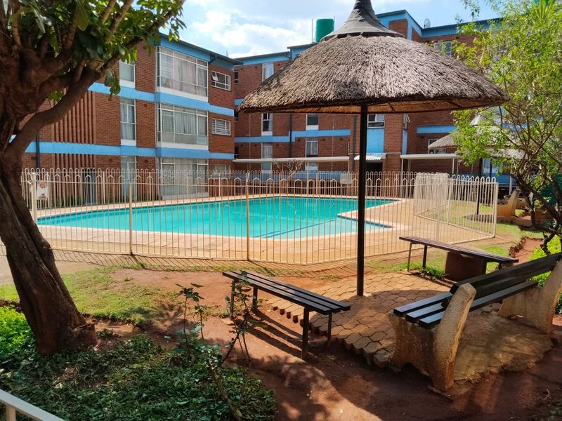Communal swimming pool area featuring a fenced pool, thatched umbrella, and paved seating area set within the apartment complex grounds.