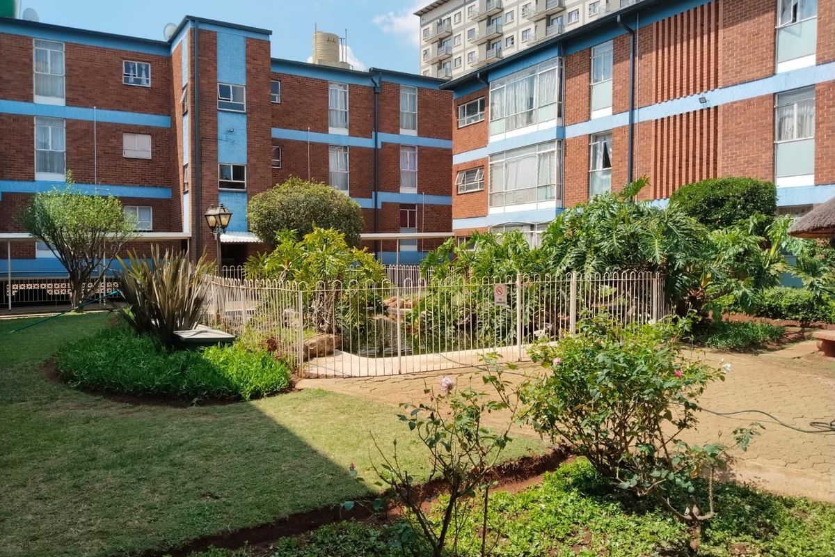 A well-maintained communal garden area featuring paved pathways, established landscaping, and a fenced water feature set between apartment building blocks.