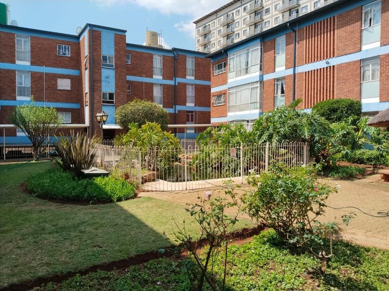 A well-maintained communal garden area featuring paved pathways, established landscaping, and a fenced water feature set between apartment building blocks.