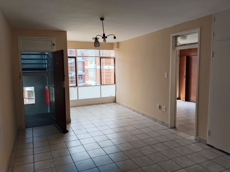 An empty, open-plan living area featuring tiled flooring, large windows for natural light, and direct access to the building's stairwell and adjacent rooms.