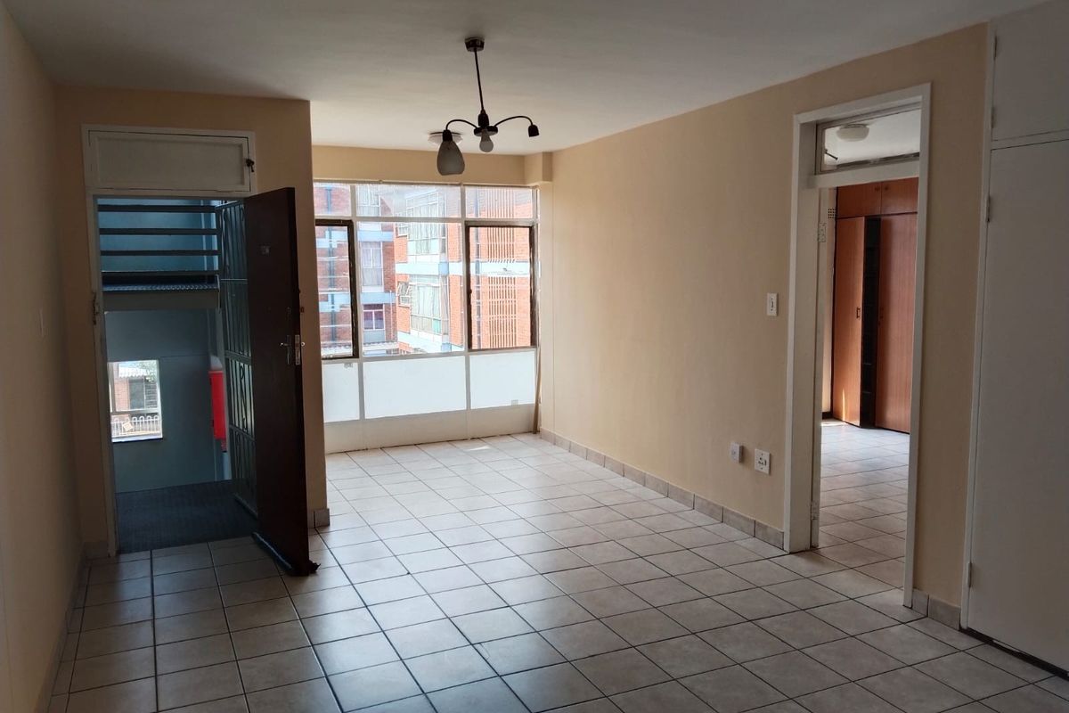 An empty, open-plan living area featuring tiled flooring, large windows for natural light, and direct access to the building's stairwell and adjacent rooms.
