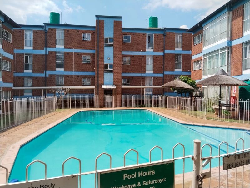 Communal swimming pool area featuring a fenced pool, paved surround, and thatched umbrellas set against a multi-story brick apartment building.