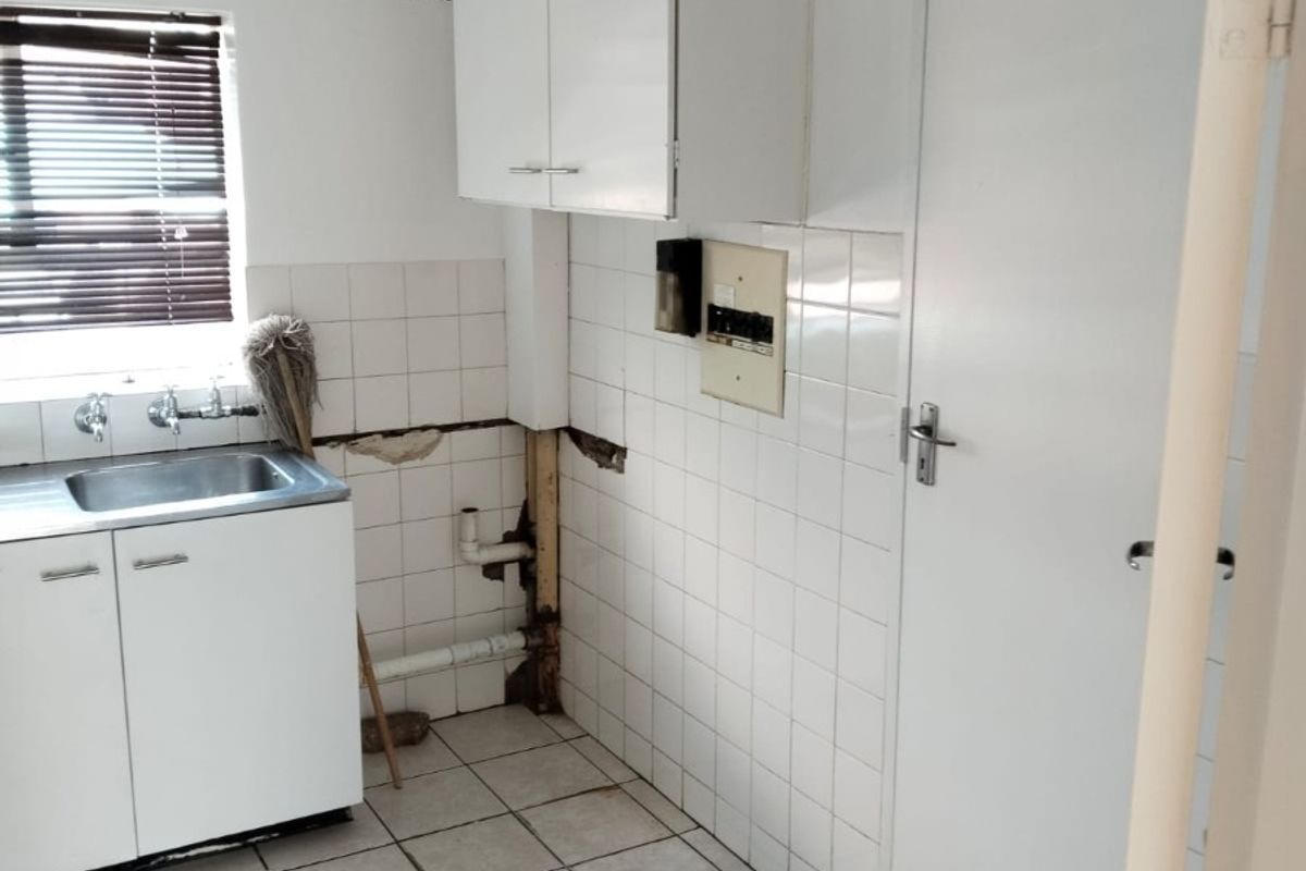 A small kitchen area featuring tiled flooring, a sink, and wall-mounted cabinetry, currently requiring repairs to the wall tiling and exposed plumbing infrastructure.
