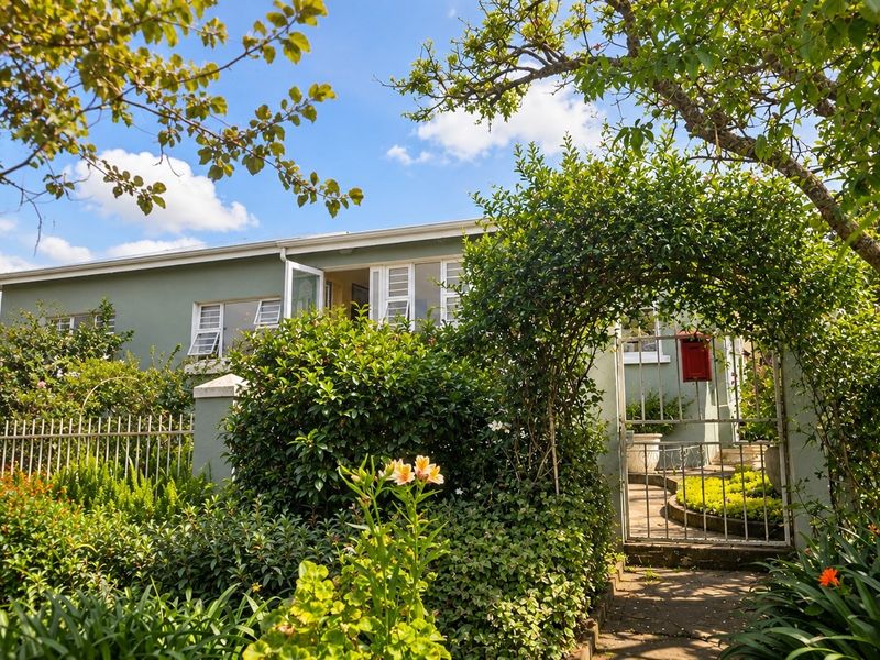 A charming front exterior view of the house featuring a gated entrance, lush garden landscaping, and a paved pathway leading to the main entrance.