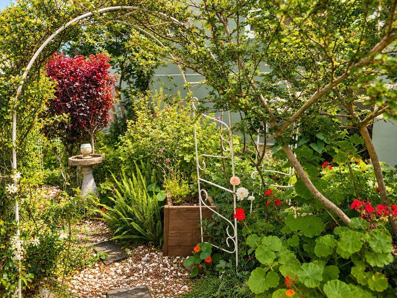 A lush, well-established garden featuring a metal archway, gravel pathways, stone pavers, and a decorative bird bath surrounded by mature greenery and flowering plants.