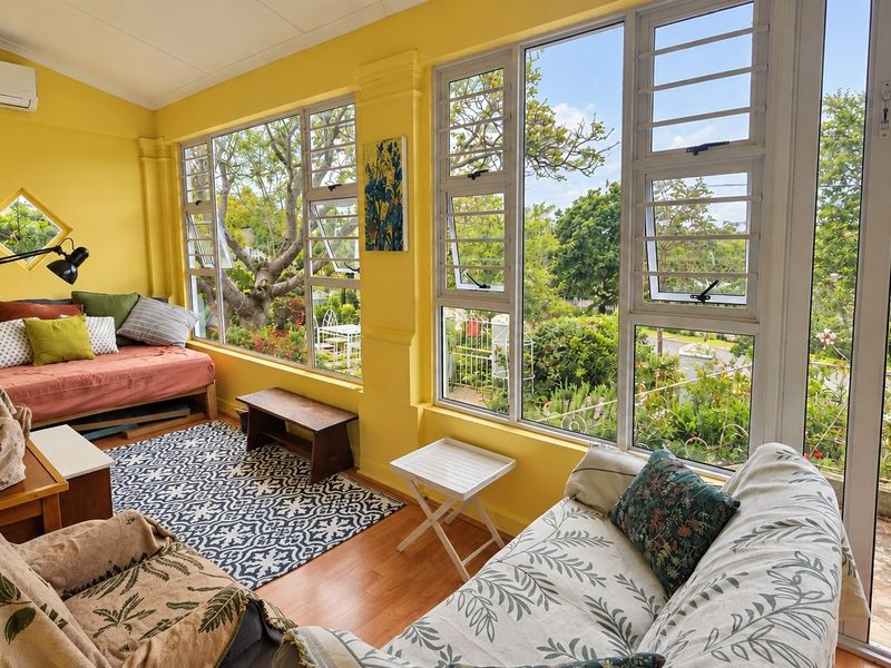 A bright, sun-filled living room featuring large windows with garden views, laminate flooring, and a wall-mounted air conditioning unit.