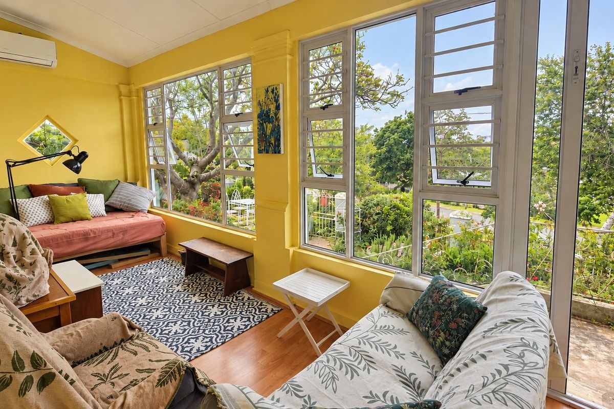 A bright, sun-filled living room featuring large windows with garden views, laminate flooring, and a wall-mounted air conditioning unit.