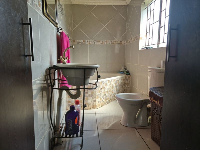 A functional bathroom featuring tiled walls and flooring, a built-in bathtub, a pedestal basin, and a window with security bars.