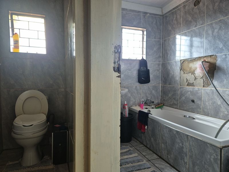 A bathroom featuring a bathtub and toilet with grey tiled finishes, currently showing signs of wear including missing wall tiles and damaged door frames.