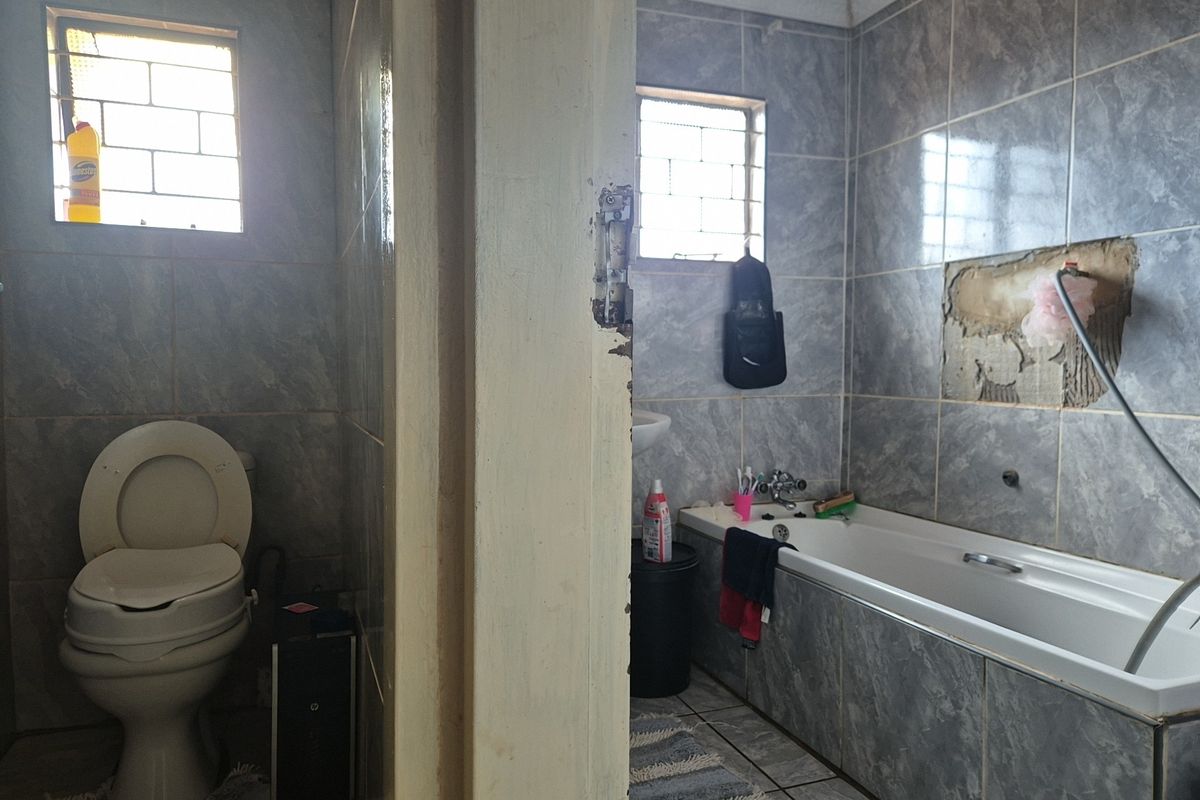 A bathroom featuring a bathtub and toilet with grey tiled finishes, currently showing signs of wear including missing wall tiles and damaged door frames.