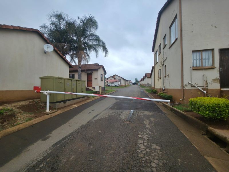 A view of the residential complex entrance featuring a security boom gate, asphalt driveway, and multiple housing units with tiled roofs.