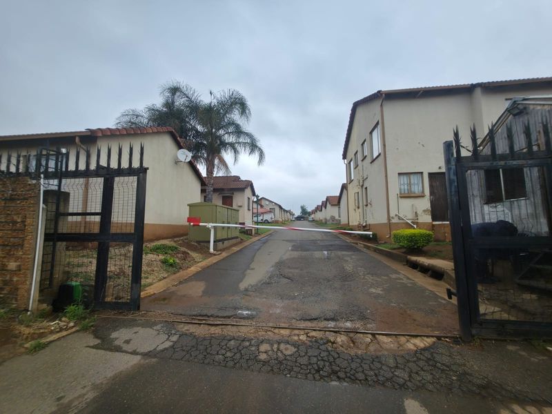 View of a residential complex entrance featuring a security gate, boom barrier, and asphalt driveway leading to multiple housing units.