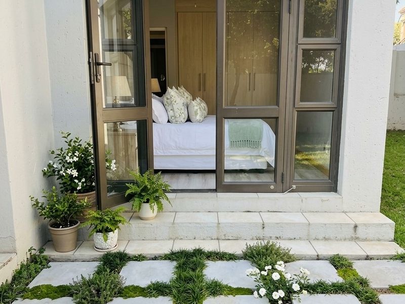 A view of a bedroom interior from a paved rear patio, featuring aluminium glass doors and a well-maintained garden setting.
