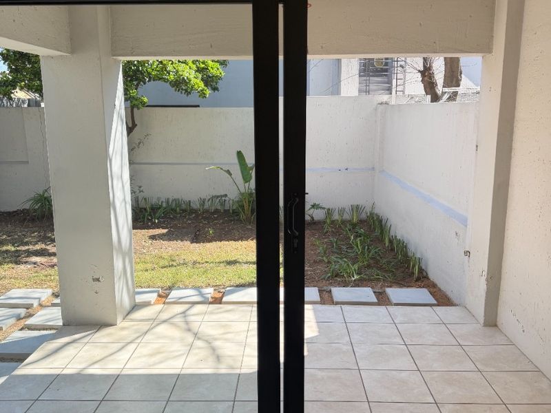 A covered patio area featuring tiled flooring and concrete pillars, opening onto a small private garden space with a boundary wall.