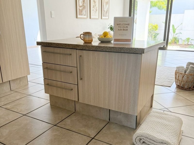A modern kitchen island featuring light wood-grain cabinetry and a stone countertop, set against a bright, open-plan living area with tiled flooring.