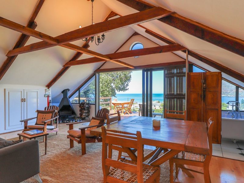 Spacious open-plan living area featuring vaulted ceilings with exposed timber beams, a freestanding fireplace, and large sliding doors opening to a scenic coastal view.