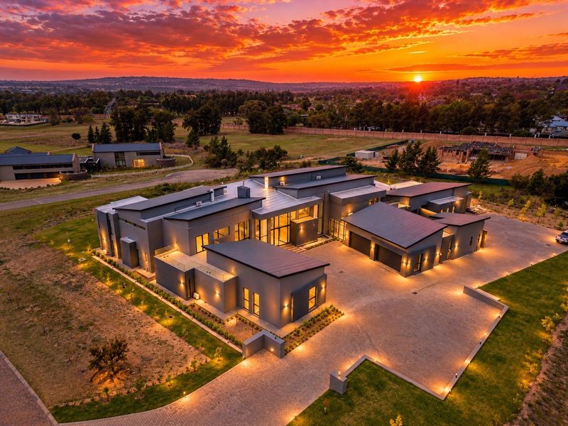 Aerial view of a modern luxury residence featuring a multi-wing architectural design, metal roofing, extensive glass facades, and a large paved driveway at sunset.